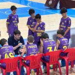 Basketball coach talks to a huddle of young players in purple uniforms on the bench during a timeout in a gym.