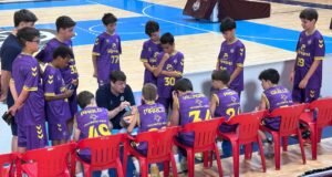 Basketball coach talks to a huddle of young players in purple uniforms on the bench during a timeout in a gym.