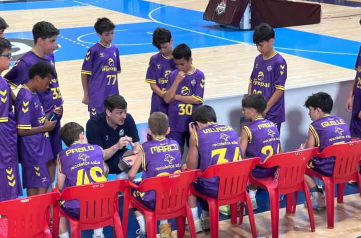 Basketball coach talks to a huddle of young players in purple uniforms on the bench during a timeout in a gym.