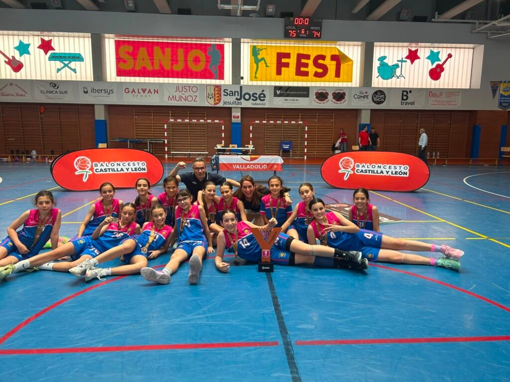 Young girls basketball team posing with medals and trophy on a gym floor, indoors with banners behind them