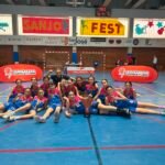 Young girls basketball team posing with medals and trophy on a gym floor, indoors with banners behind them