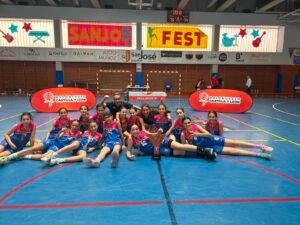 Young girls basketball team posing with medals and trophy on a gym floor, indoors with banners behind them