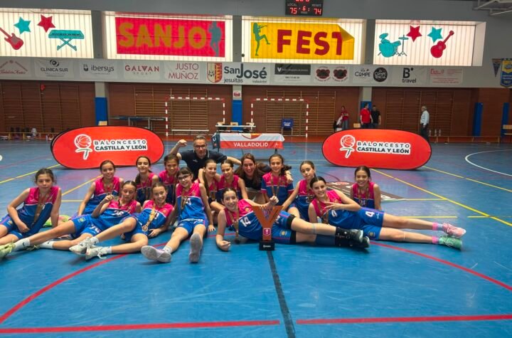 Young girls basketball team posing with medals and trophy on a gym floor, indoors with banners behind them