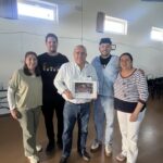 Five adults pose together indoors, with the man in a white shirt holding a framed photo or certificate in the center.