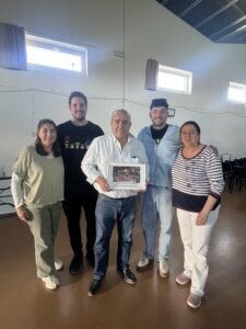 Five adults pose together indoors, with the man in a white shirt holding a framed photo or certificate in the center.