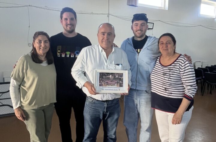Five adults pose together indoors, with the man in a white shirt holding a framed photo or certificate in the center.