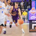 Basketball player in a purple uniform dribbles past white-uniform defenders during a fast break on a polished court with spectators in the background.