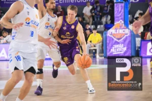 Basketball player in a purple uniform dribbles past white-uniform defenders during a fast break on a polished court with spectators in the background.