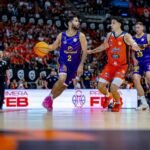 Basketball players in purple and orange uniforms compete for the ball on a crowded indoor court with a cheering crowd in the background.
