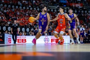 Basketball players in purple and orange uniforms compete for the ball on a crowded indoor court with a cheering crowd in the background.