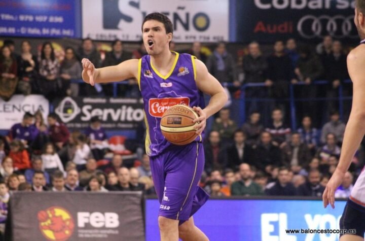 Basketball player in purple jersey shouts while dribbling the ball on court, with a packed crowd in the background.