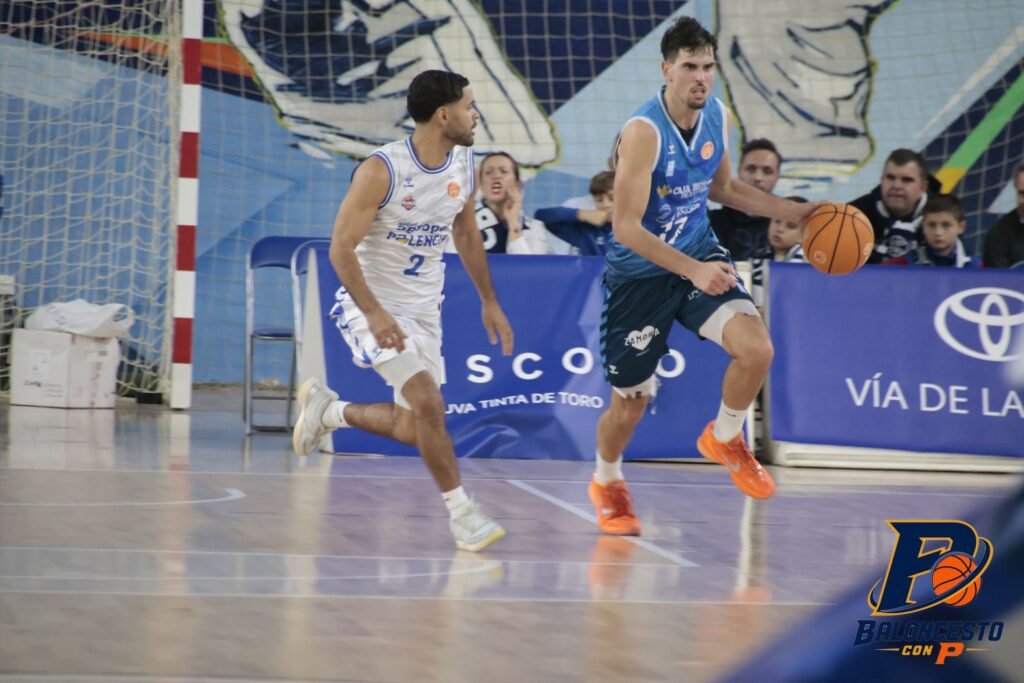 Basketball players in white and blue jerseys chase the ball on an indoor court, with orange shoes and a blue advertising banner in the background.