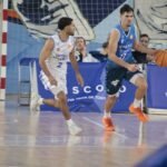 Basketball players in white and blue jerseys chase the ball on an indoor court, with orange shoes and a blue advertising banner in the background.