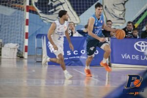 Basketball players in white and blue jerseys chase the ball on an indoor court, with orange shoes and a blue advertising banner in the background.