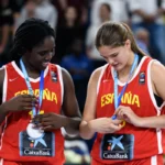 Two young female players in red España basketball jerseys smile as they wear medals at a podium ceremony; CaixaBank logos visible on their uniforms.