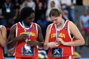 Two young female players in red España basketball jerseys smile as they wear medals at a podium ceremony; CaixaBank logos visible on their uniforms.