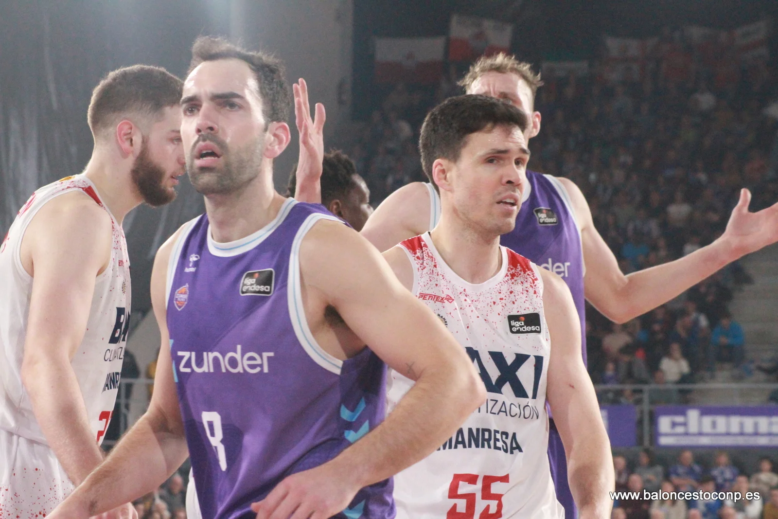 Group of male basketball players on the court during a game, wearing purple and white uniforms, with a crowded arena in the background.