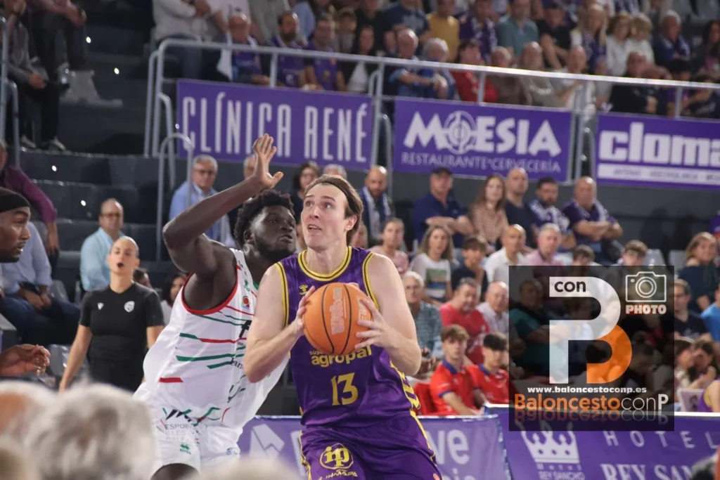 Basketball player in purple (#13) holds the ball while a defender in white blocks her, in a crowded arena with banners behind them.