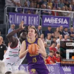 Basketball player in purple (#13) holds the ball while a defender in white blocks her, in a crowded arena with banners behind them.