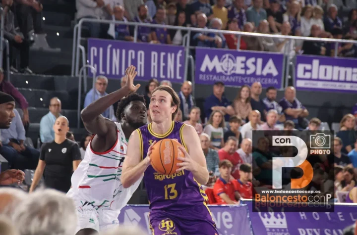 Basketball player in purple (#13) holds the ball while a defender in white blocks her, in a crowded arena with banners behind them.