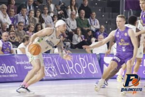 Basketball player in white drives with the ball while defending in a crowded arena.