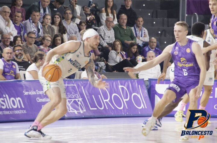 Basketball player in white drives with the ball while defending in a crowded arena.