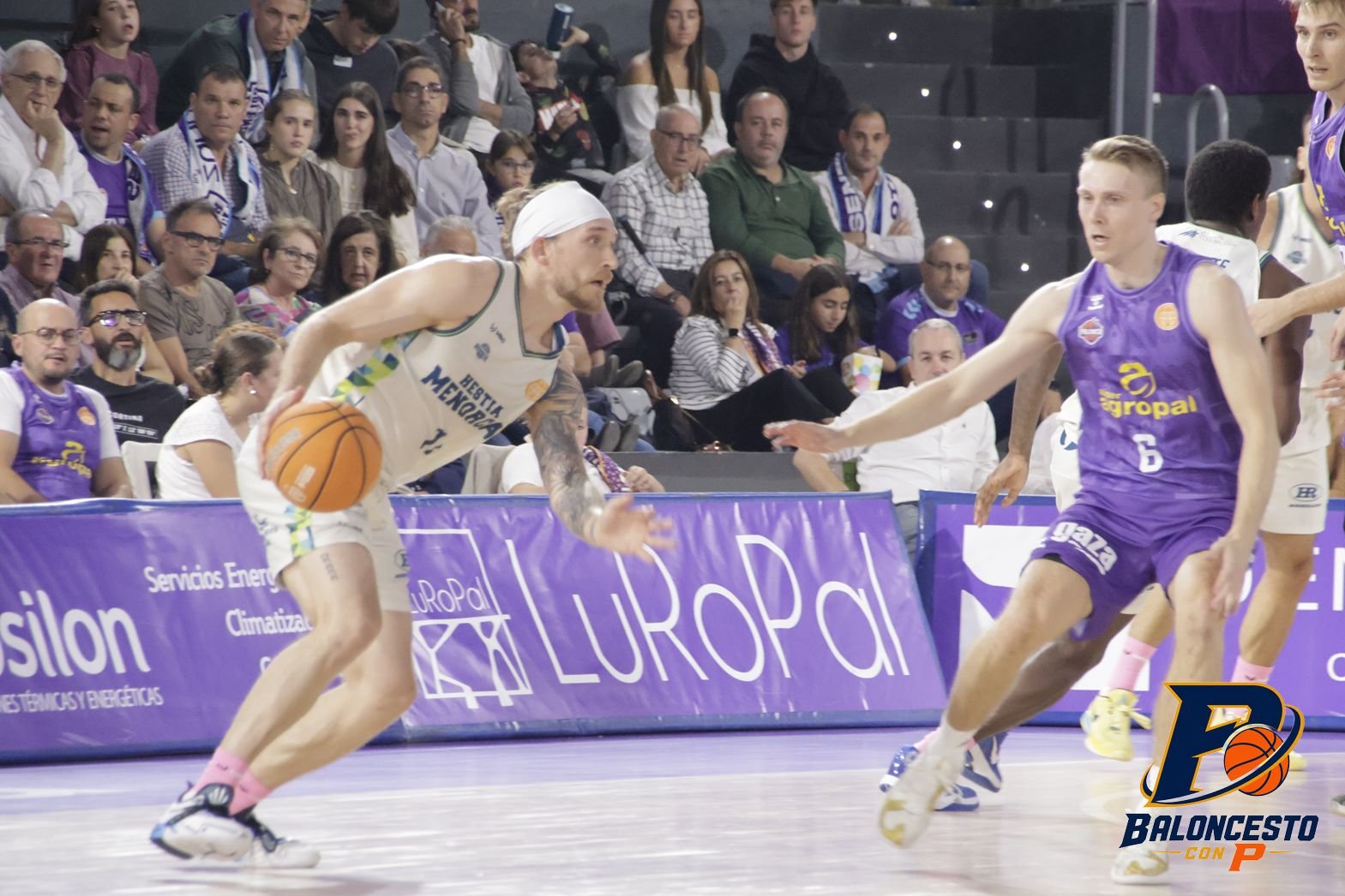 Basketball player in white drives with the ball while defending in a crowded arena.