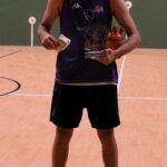 Male basketball player standing on an indoor court, wearing a purple jersey and black shorts, holding a trophy in one hand and a card in the other, with a bottle on the floor at his feet.