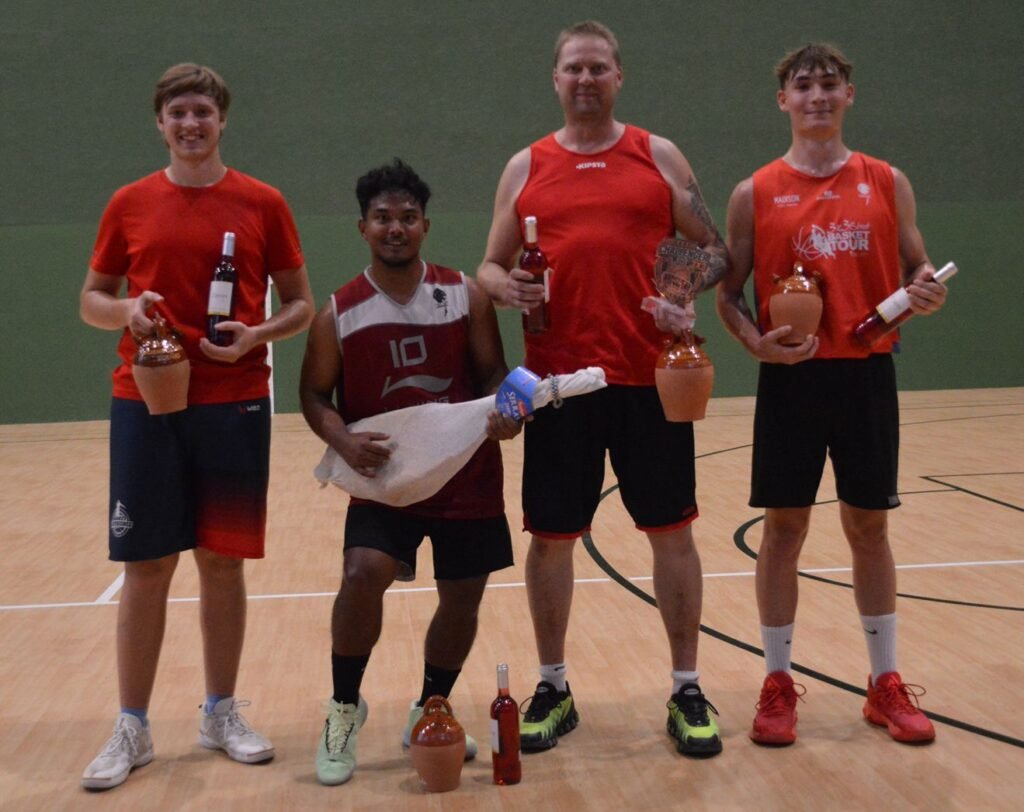 Five basketball players stand on a gym floor posing with trophies, bottles, and prizes after a game (center-right holding a plaque).