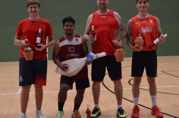 Five basketball players stand on a gym floor posing with trophies, bottles, and prizes after a game (center-right holding a plaque).