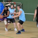 Four players compete indoors on a wooden basketball court; a black‑clad player guards the ball while two blue‑shirted players defend near the hoop.