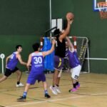 Basketball players in purple jerseys contest a shot near the hoop inside a gymnasium, with one defender jumping to block.”,