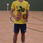 Young man in a yellow shirt and blue shorts standing on an indoor basketball court, holding a trophy and a bottle.