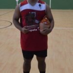 Male athlete in a red jersey stands on an indoor court, holding an orange ceramic jug trophy with a bottle on the floor between his feet.
