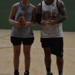 Woman and man in gym clothes stand side by side on a basketball court, each holding a brown trophy vase; two red bottles sit on the floor before them.