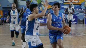 Basketball player in blue drives with the ball toward the basket while defenders in white surround him on an indoor court with a colorful mural backdrop.