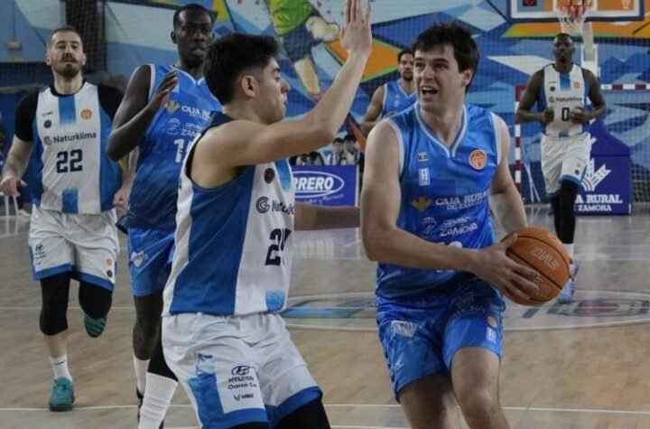 Basketball player in blue drives with the ball toward the basket while defenders in white surround him on an indoor court with a colorful mural backdrop.