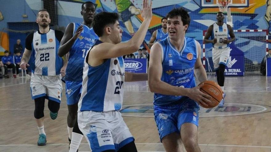 Basketball player in blue drives with the ball toward the basket while defenders in white surround him on an indoor court with a colorful mural backdrop.