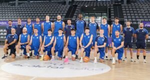 Basketball team photo: players kneeling in front, standing behind in blue uniforms on a gym court with empty bleachers in background