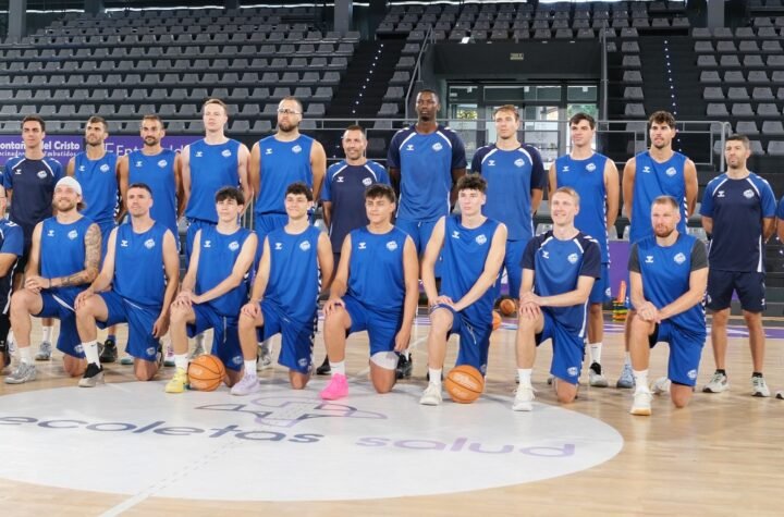 Basketball team photo: players kneeling in front, standing behind in blue uniforms on a gym court with empty bleachers in background