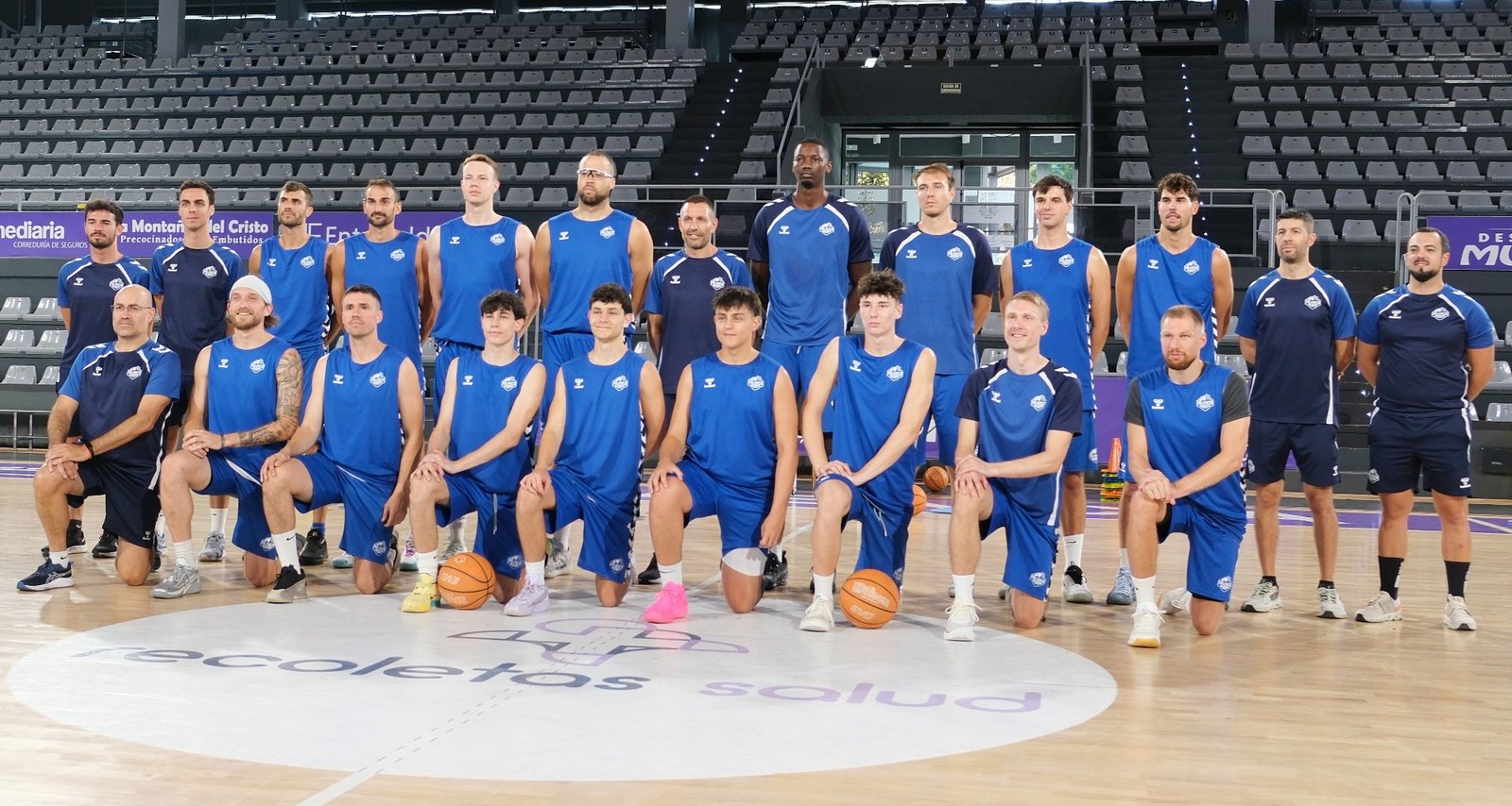 Basketball team photo: players kneeling in front, standing behind in blue uniforms on a gym court with empty bleachers in background